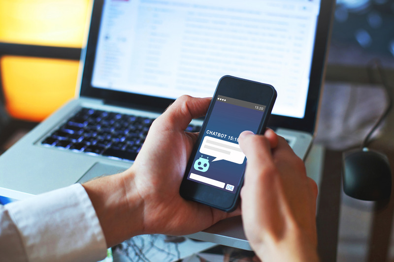 a-person-using-a-cell-phone-in-front-of-a-laptop-computer-the-person-is-holding-the-cell-phone-in-their-right-hand-and-is-looking-at-the-screen-the-laptop-is-open-and-is-displaying-a-website-with-the-text-chatbot-15-1-there-is-also-a-region-of-the-screen-that-shows-the-antares-chatbot-logo-the-image-was-taken-in-a-well-lit-room-and-the-background-is-mostly-white
