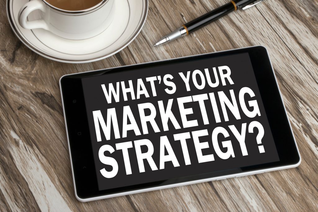 a-tablet-with-the-words-whats-your-marketing-strategy-written-on-it-the-tablet-is-sitting-on-a-wooden-table-next-to-a-cup-of-coffee-and-a-pen