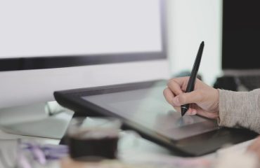 virtual-graphic-designer-working-on-a-digital-pad-with-a-digital-pen-using-imac-in-the-background-while-enjoying-a-cup-of-coffee