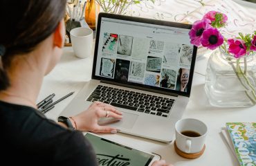 virtual-graphic-designer-working-on-imac-using-pinterest-for-inspiration-typing-on-keyboard-with-coffee-and-plants-in-the-background-an-ipad-and-sketchpad-with-pens-are-also-visible-on-the-desk